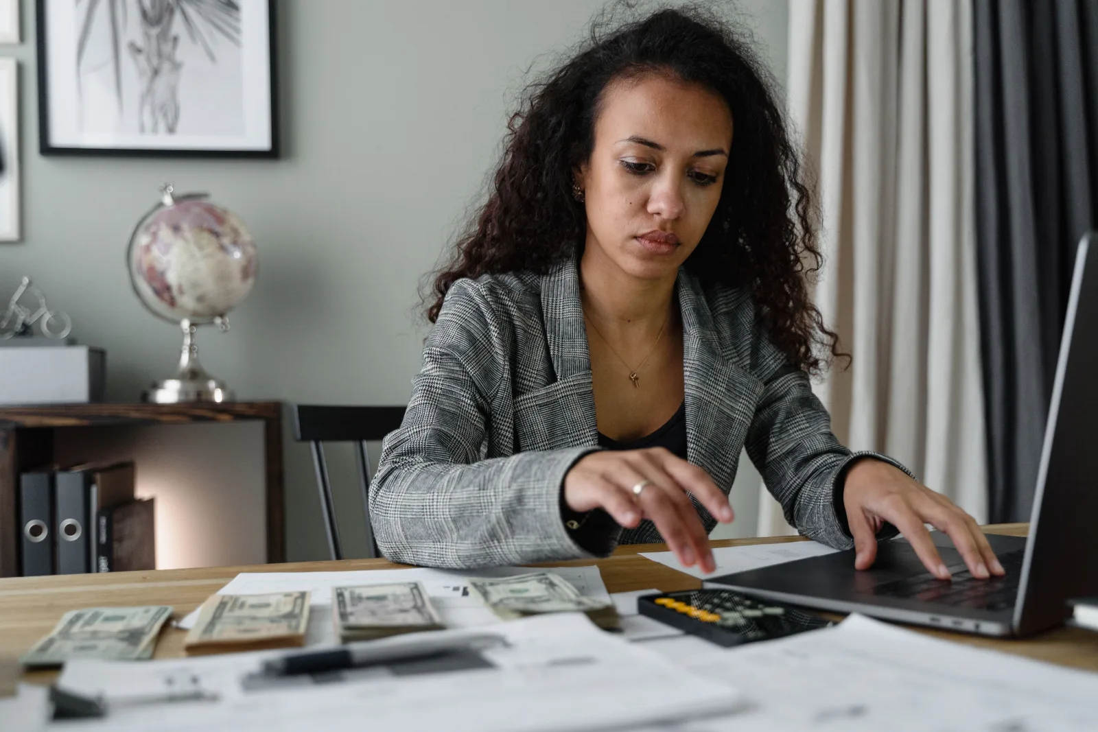 An African woman reviews her finances at a desk, using a laptop and calculator while examining cash, bills, and financial documents.