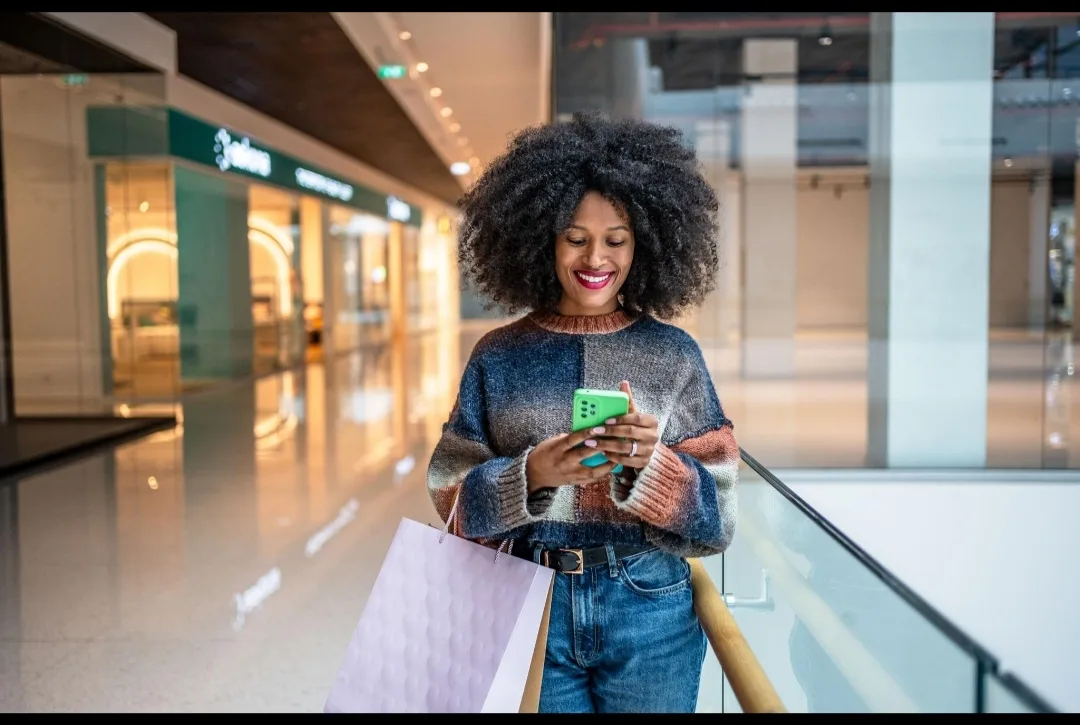 A young African woman carries shopping bags while using her phone, illustrating lifestyle spending.